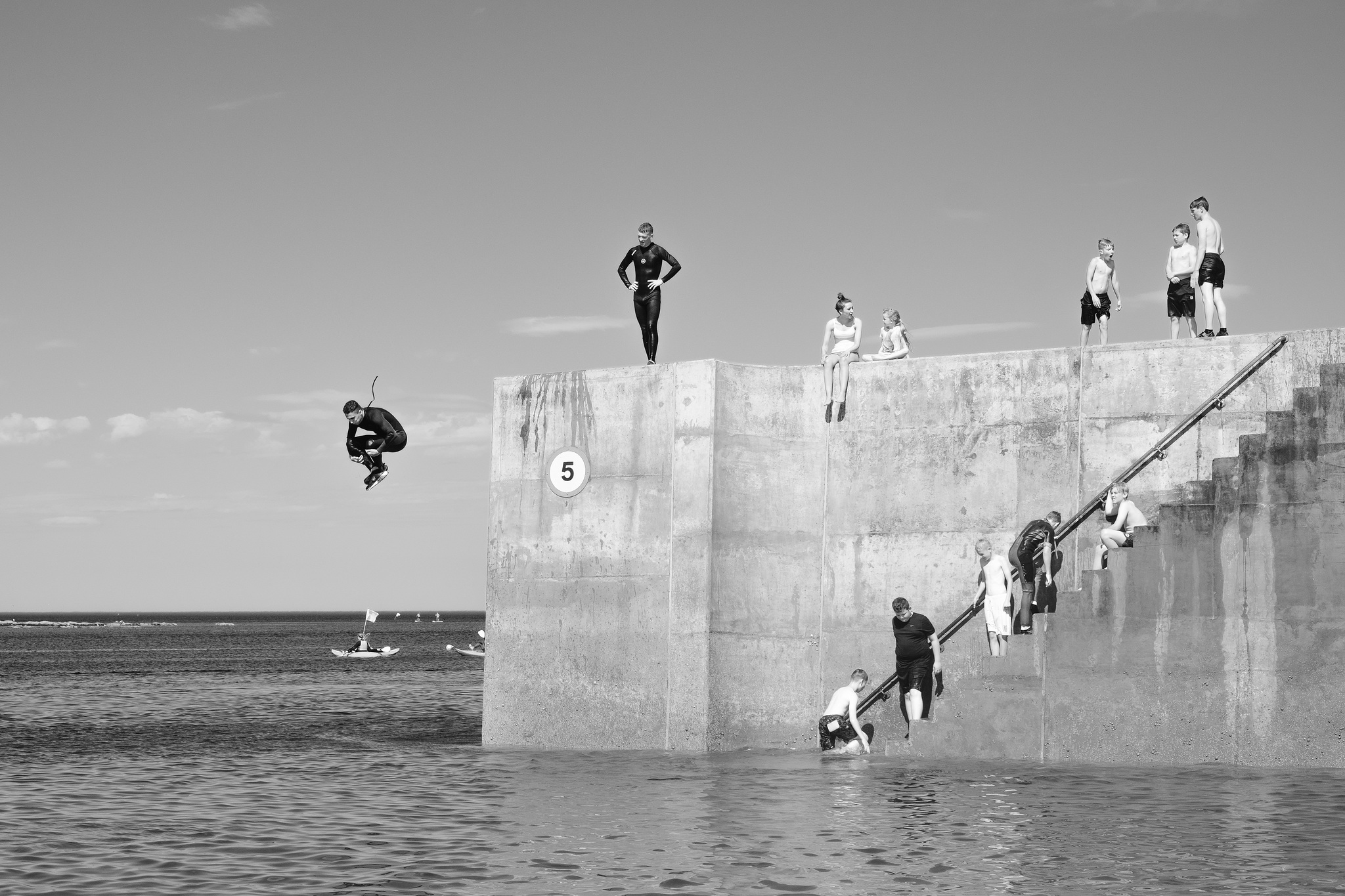 Teenage boy in wetsuit jumps off a harbour wall into the water while groups of other kids wait on the wall and steps Teenage boy in wetsuit jumps off a harbour wall into the water while groups of other kids wait on the wall and steps
